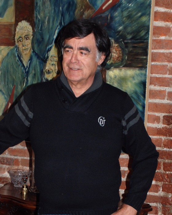 Portrait de Floréal Plana : un homme aux cheveux bruns, en pull noir, debout devant un mur de briques et une toile peinte.